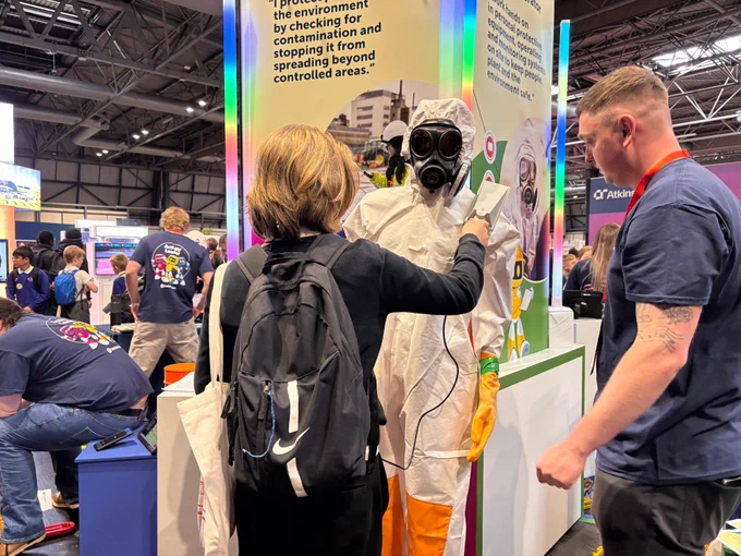 A mannequin wearing a hazmat suit and gas mask is having their badge scanned at The Big Bang Fair. Another person in casual attire is assisting with the scanning process. The event appears lively, with various booths and attendees in the background.