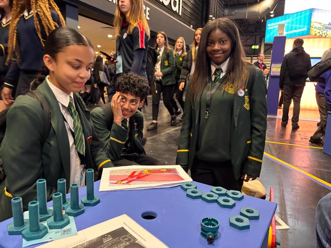 A group of students in school uniforms is gathered around a table displaying a model and large nuts and bolts. The students are engaged in conversation, with one looking at the camera and the others focused on the table. 