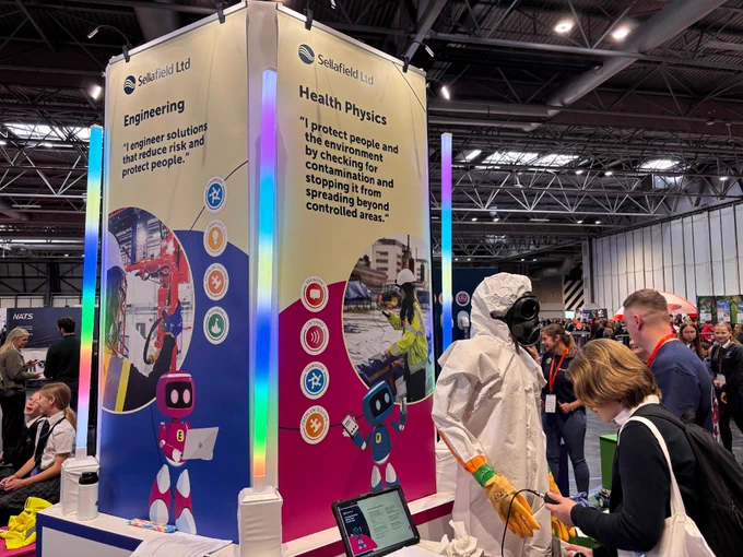 A bustling exhibition booth for Sellafield Ltd displays two vertical signs titled "Engineering" and "Health Physics," each with colorful graphics. The booth includes a mannequin wearing a protective suit and a visitor engaging with a staff member. Bright lights and additional stalls are visible in the background.