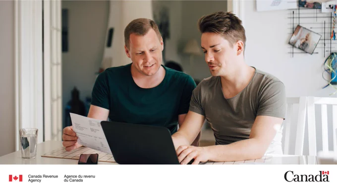 Two people sit at a kitchen table while looking at a laptop and a printed document.
