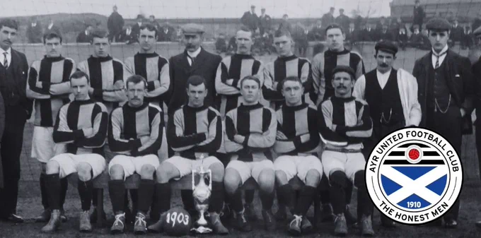 Black-and-white historic team photograph of early Ayr United football players, arranged in two rows on a pitch. The front row is seated with a trophy placed on the ground in front, marked “1903,” while the back row stands behind. The players wear striped football kits with long socks, and several spectators are visible in the background. An Ayr United Football Club crest is overlaid in the bottom right corner.