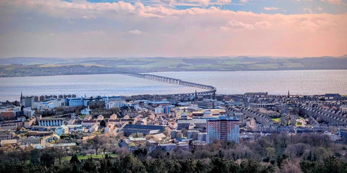 A blue, cloudy sky above a city, a bridge over a river and landscape in the distance.