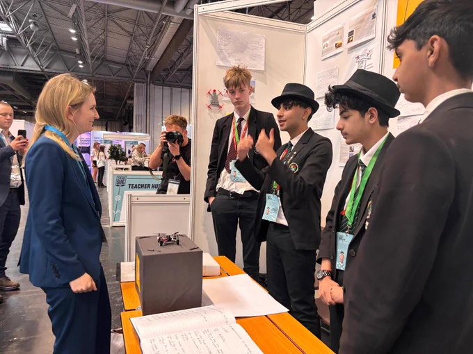 Shadow SoS for Science, Innovation & Technology Julia Lopez MP,  is speaking with a group of individuals dressed in school uniforms at The Big Bang Fair. They are Big Bang Competition finalists. The booth features a project displayed on the table, with drawings and documents on the board behind them. People are walking in the background, and an individual is taking photos.