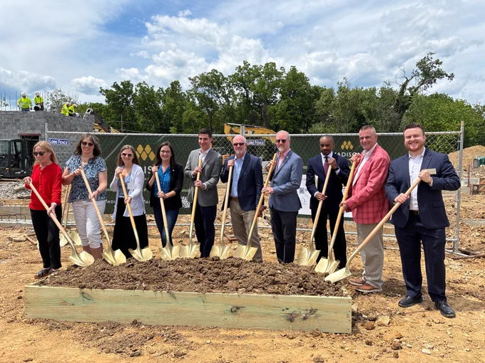 Several men and women, dressed in business attire, pose with shovels in the dirt to celebrate the groundbreaking of a new affordable housing development.
