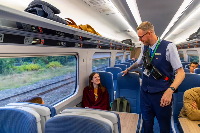 A TransPennine Express train conductor interacts with a passenger inside a train carriage with blue seats and luggage overhead.