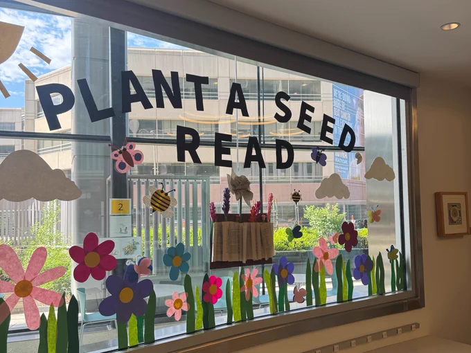 Colorful paper decorations adorn a large window. The window features the words "PLANT A SEED READ" in bold black letters. Below, vibrant paper flowers in various colors, alongside bees, butterflies, and clouds, create a lively scene. A book is open and displayed in the center of the arrangement. The background shows an urban building and a bright blue sky.