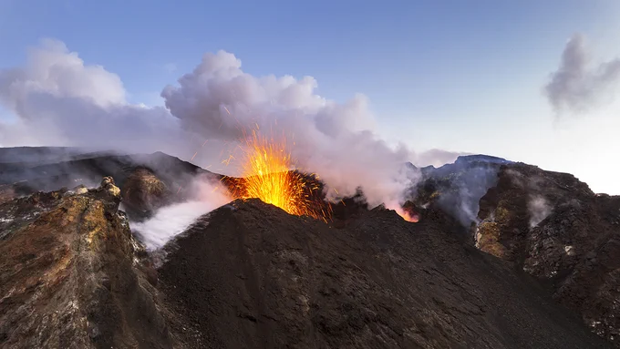 A vibrant volcanic eruption with molten lava and bright sparks rises from a crater, surrounded by rocky terrain and clouds of smoke, set against a serene blue sky.