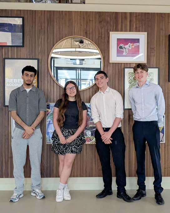 Four people standing in front of a wooden wall with framed artwork and a circular mirror in an office setting.