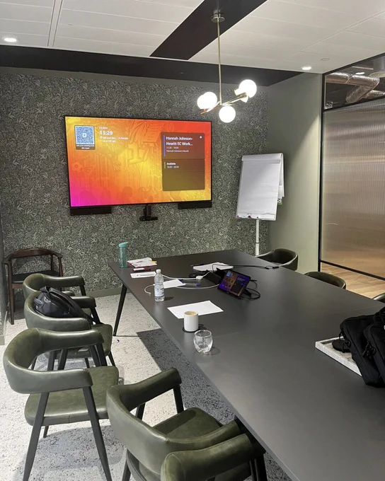 Modern meeting room with a long table, green chairs, wall-mounted screen displaying a meeting schedule, and flip chart to the side.