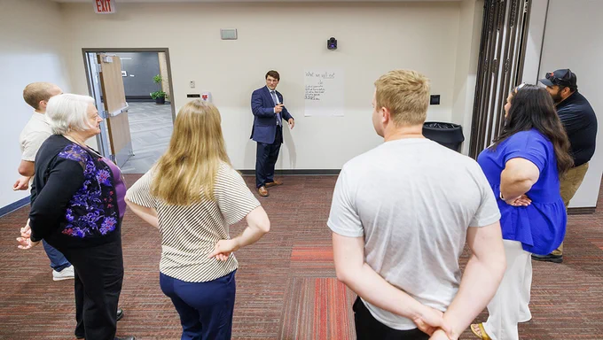 
Timothy "Ben" Bentzinger (center), wearing a dark blue suit with a tie, takes notes on a large piece of paper at the front of a big room, as three men and three women stand in front of him in a semicircle. (Jordan Opp/University Communication and Marketing)