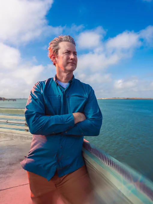 A man stands with arms folded next to a short wall, looking out at the ocean. Photo credit: Tico Mendoza