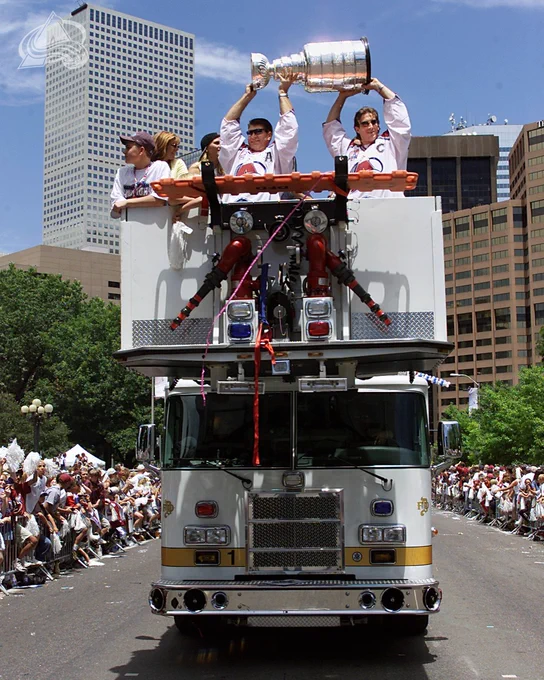 Joe Sakic and Raymond Bourque lifting the cup over their head on top of a fire truck as it drives through the streets of Denver