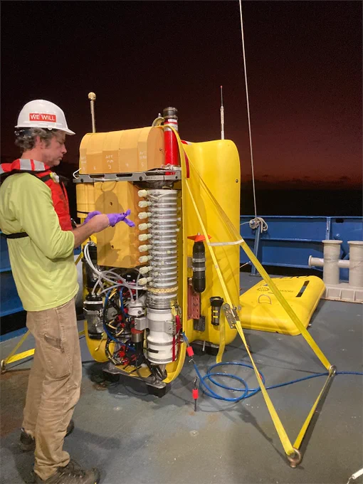 A man stands on the deck of a research vessel, working on a robotic submersible that looks like a large yellow box about the size of a small car. Photo credit: Chip Breier.