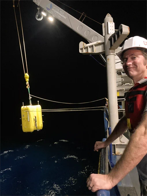 A man leans against a railing on the deck of a research vessel. Behind him, a crane is lowering a robotic submersible that looks like a large yellow box about the size of a small car into the water. Photo credit: Chip Breier