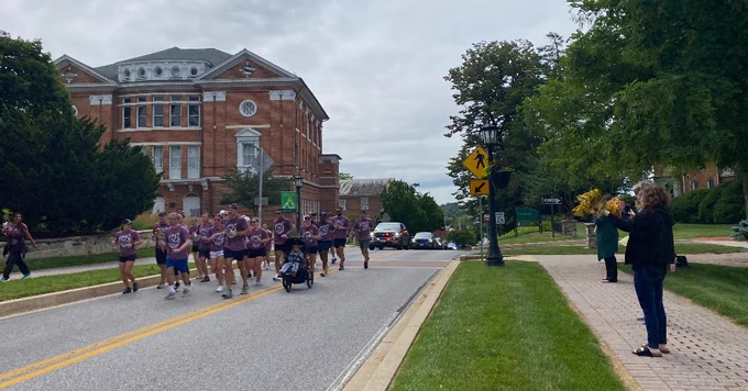A person on a motorcycle leads a group of people in matching purple shirts as they jog down a road. On the sidewalk, a few onlookers cheer them on, some holding pompoms. A large brick building is visible in the background, surrounded by trees and greenery.