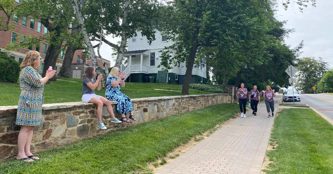 A group of individuals are walking on a sidewalk, while several others are sitting on a stone wall nearby cheering. 