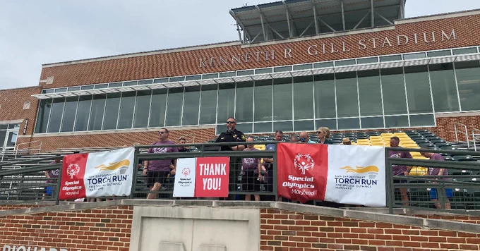 McDaniel College Director of Campus Safety Eric Immler speaks to a group of individuals standing in a stadium with Special Olympics Maryland banners displayed.  The backdrop includes brickwork and stadium seating.