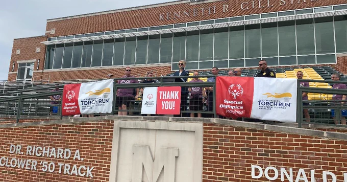 McDaniel College President Julia Jasken speaks to a group of individuals standing in a stadium with Special Olympics Maryland banners displayed.  The backdrop includes brickwork and stadium seating.