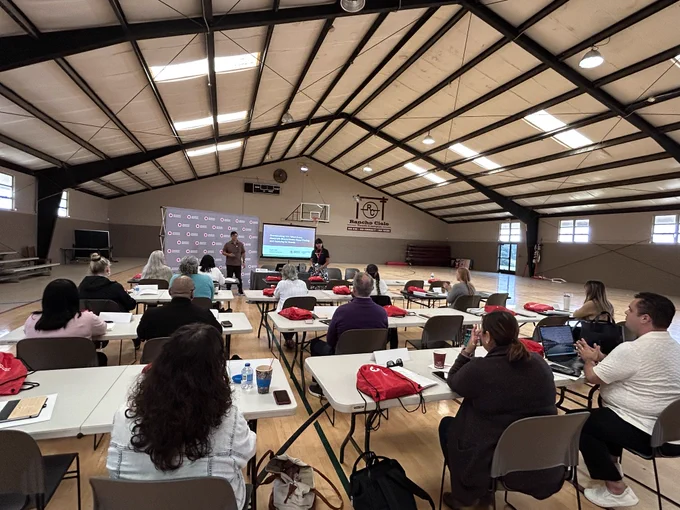 A group of people seated at tables in a gymnasium attend a presentation. There is a speaker at the front with a screen displaying information and a backdrop with the American Red Cross logo.