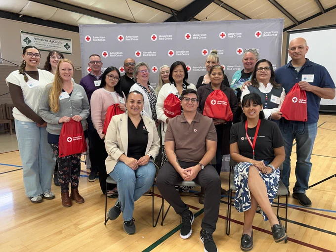 A group of people posing in front of an American Red Cross backdrop inside a gymnasium. Some are holding Red Cross gift bags, and various individuals are dressed in casual and business attire. American Red Cross logos are visible on the backdrop and bags.