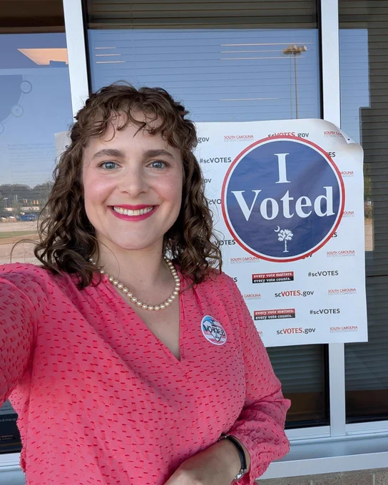 Mary Alex, a white woman in a coral top smiles with I voted sticker and sign at her polling location.