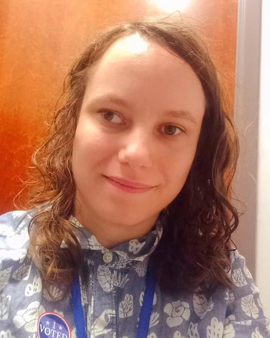 Julia, a white woman with curly brown hair using a wheelchair, takes a selfie with her 'I voted' sticker.