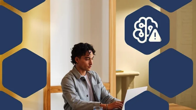Person sitting at a desk working on a laptop, surrounded by blue hexagon shapes with an AI brain warning icon, representing AI-related security risks and social engineering threats.