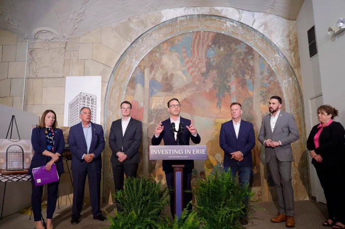Governor Josh Shapiro speaking from a podium with 'Investing in Erie' sign during a formal announcement in an ornate room with a group of five officials standing behind him.