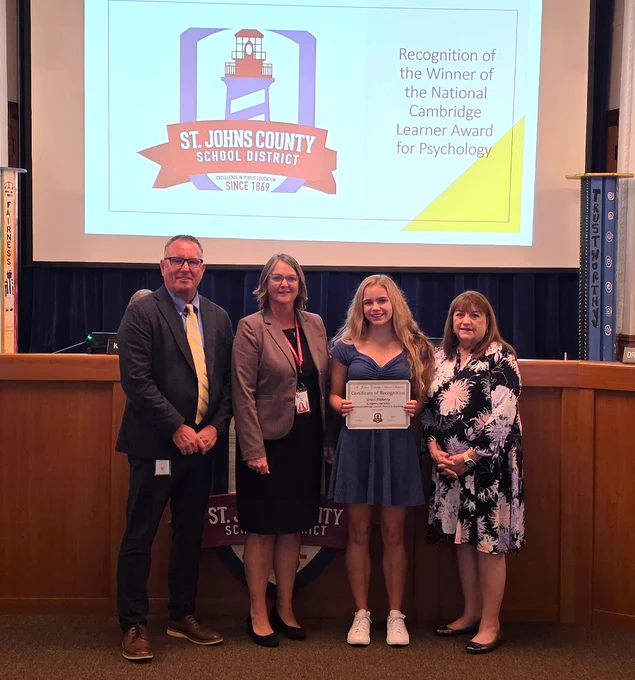 Four individuals in the St. Johns County School District auditorium with a student holding a certificate for the National Cambridge Learner Award in Psychology.