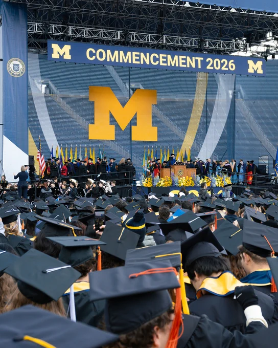 A crowd of graduates looks toward the stage during the University of Michigan's 2026 Spring Commencement ceremony in Michigan Stadium. A large maize Block M prominently hangs in the background.