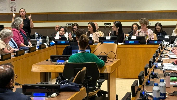 UN conference room where participants sit with laptops and microphones. Several speakers are seated at the front, with nameplates in front of them. A sign language interpreter stands nearby, raising both hands while signing. Water bottles and documents are placed on tables.