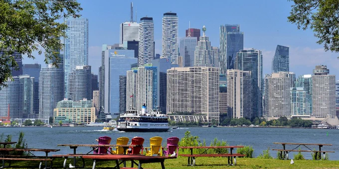 Colourful chairs and picnic tables on a grassy park overlooking a ferry crossing a river with a city skyline of tall modern buildings in the background.