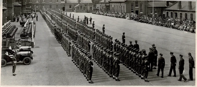 A large military parade is taking place in a wide courtyard surrounded by brick buildings. Numerous uniformed persons stand in organized rows. Onlookers are seated along the periphery. Several jeeps are parked to the side. The scene captures a formal gathering with a sense of order and discipline.