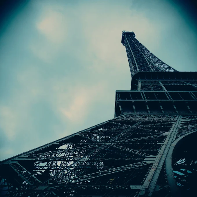 Low-angle photograph of the Eiffel Tower in Paris, rising diagonally through a pale blue sky. The iron structure fills the frame with dark steel lines and geometric detail, giving the image a calm, monumental, and atmospheric presence.