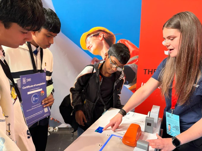 A group of individuals is gathered around a table at a tech exhibit. One individual is demonstrating a device while others observe attentively, holding certificates. In the background, a large image shows a person in construction gear.