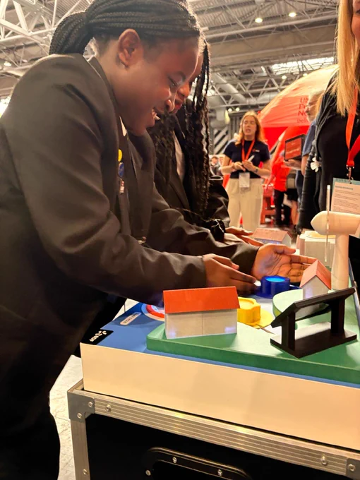 A person smiles while interacting with a table featuring small models of colorful buildings. Other people are present in the background at what appears to be an event or exhibition.