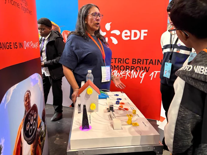 An individual is presenting an interactive display at an EDF booth. The display features a model house and various energy components. Several people are engaged and listening attentively. EDF branding is clearly visible on a red backdrop.