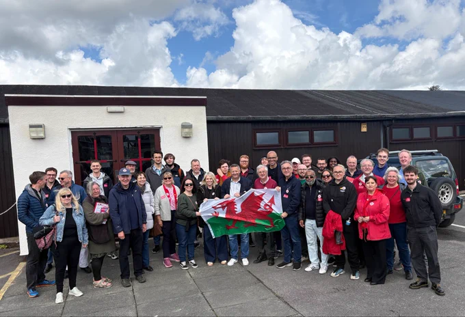 A photo of Labour members stood in front of a building. The members at the front hold a Welsh flag.