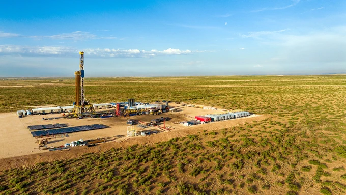 Aerial view of a large oil drilling rig in a vast desert landscape. The site is surrounded by equipment and vehicles, with a clear blue sky above.