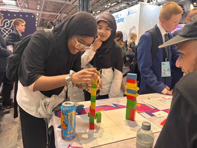 Two people are stacking colorful Jenga blocks on a table at an event. A can of Juicy Fuel Cola and a water bottle are nearby. Other attendees, including someone with a badge, are engaged around the table.