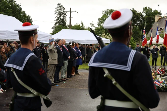 Cérémonie commémorative en extérieur sous des tentes blanches, avec des militaires en uniforme marine au premier plan, des officiels en écharpe tricolore et des civils debout en arrière-plan. Plusieurs drapeaux français et couronnes de fleurs sont visibles à droite de l'image, suggérant un hommage aux anciens combattants ou victimes de guerre.
