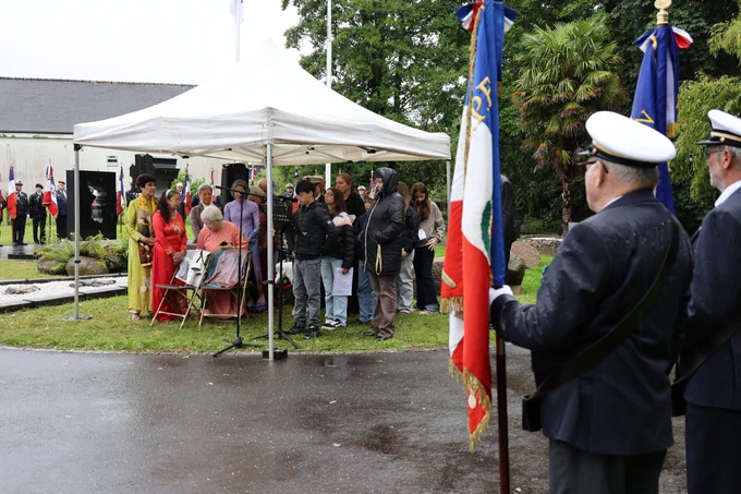 Cérémonie commémorative sous une tente blanche par temps pluvieux. Un groupe de personnes, dont certaines portent des tenues traditionnelles colorées, se tient sous la tente, tandis que des adolescents en manteaux noirs se tiennent à l’extérieur. À droite, des anciens combattants en uniforme tiennent des drapeaux tricolores français. À l’arrière-plan, une rangée de porte-drapeaux et d’autres participants se tient près d’un bâtiment clair. Décor de jardin avec arbres et pelouse humide.
