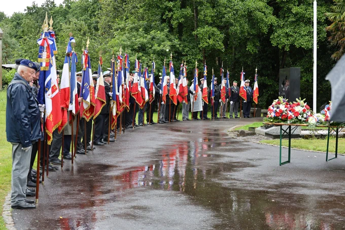 Une cérémonie commémorative en extérieur sous la pluie, rassemblant de nombreux porte-drapeaux alignés en arc de cercle, chacun tenant un drapeau tricolore français décoré de rubans. Un groupe de personnes en tenue formelle se tient devant un monument commémoratif en granit noir. Au premier plan, une table est couverte de gerbes de fleurs rouges, blanches et bleues. La scène se déroule sur une allée mouillée, entourée de verdure.