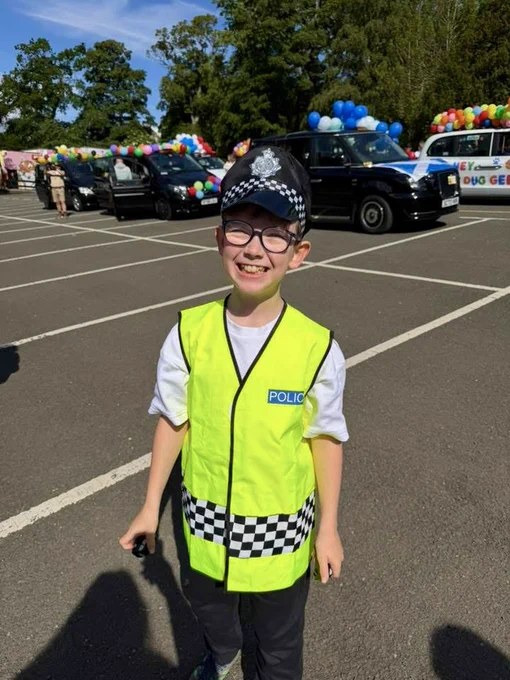 A smiling boy in a police costume, wearing a high-visibility vest and cap, stands in a car park with decorated cars in the background.