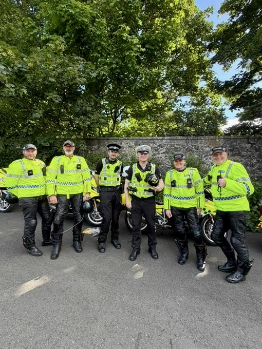 Six police officers in high-visibility jackets and motorcycle gear stand with their police motorcycles.