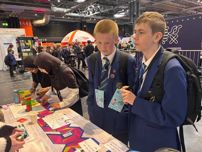 Two individuals in blue school uniforms are engaging with a display table, featuring promotional materials and colorful blocks, at an event. Other attendees and booths can be seen in the background.
