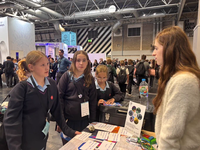 A group of individuals at an indoor event, standing at a booth. Three individuals are on one side, engaged in conversation with a person on the opposite side. The booth features informational material, a water bottle, and displays with colorful graphics. The event space is filled with people and exhibits in the background.