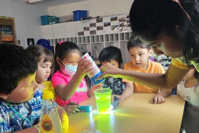 IMAGE: Photo of a group of young kids around a table in a school room, on which a luminous science experiment is being conducted.