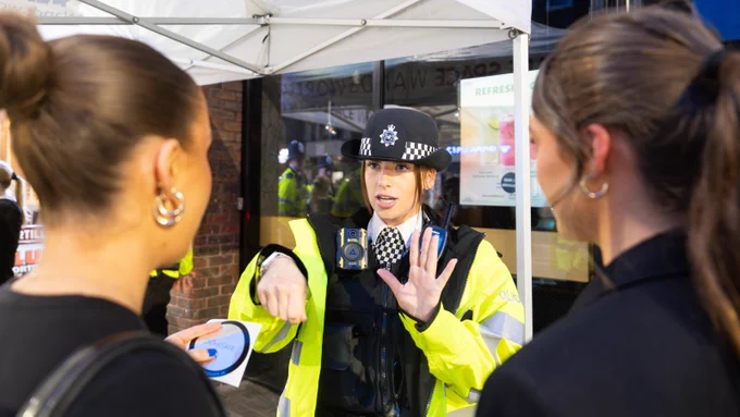 A female police officer in uniform speaks to two civilians under a tent, gesturing with her hands.