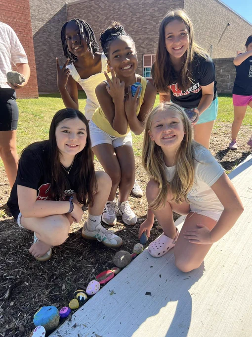 Group of five young women posing outdoors near a row of colorful painted rocks on the ground beside a sidewalk.